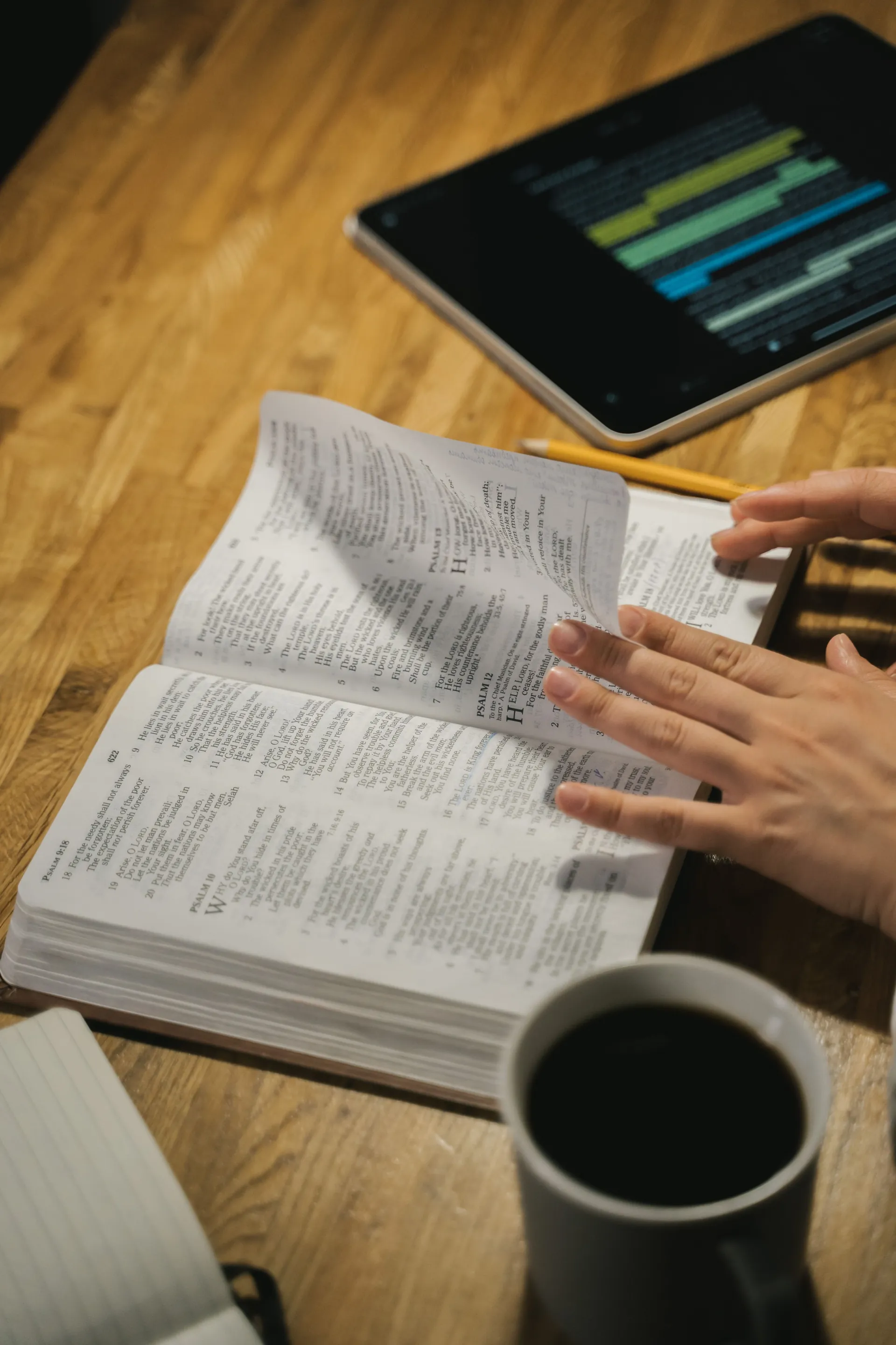 Hands turning the pages of an open Bible beside a tablet, notebook, pencil, and cup of coffee during Bible study.