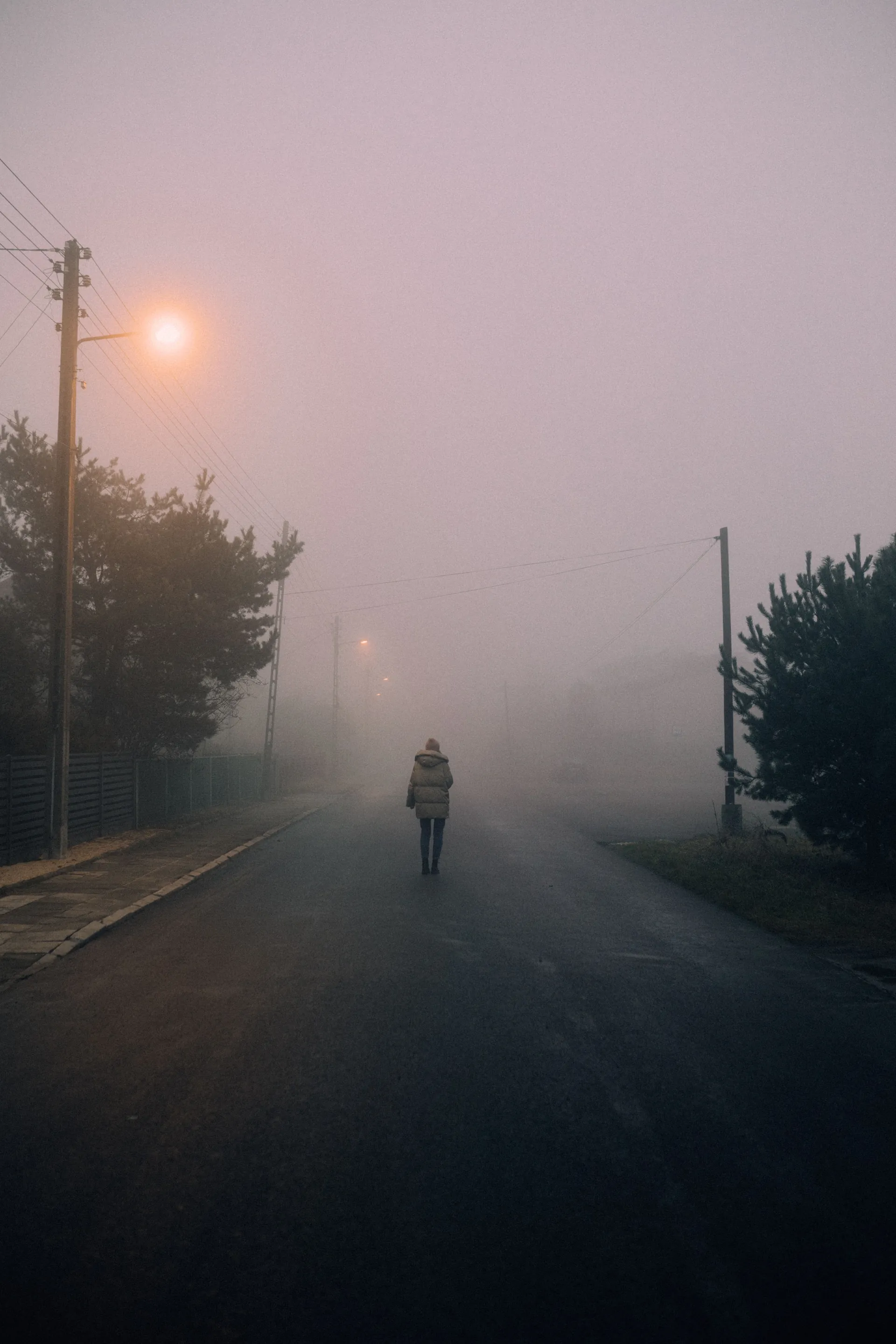 A lone person walking down a foggy street at dawn, surrounded by mist and soft streetlights.